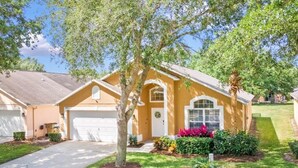 A two-story house with a garage, surrounded by well-maintained lawns and a variety of plants. - Sunshine Villa 5 Bedroom Villa by RedAwning (Kissimmee)