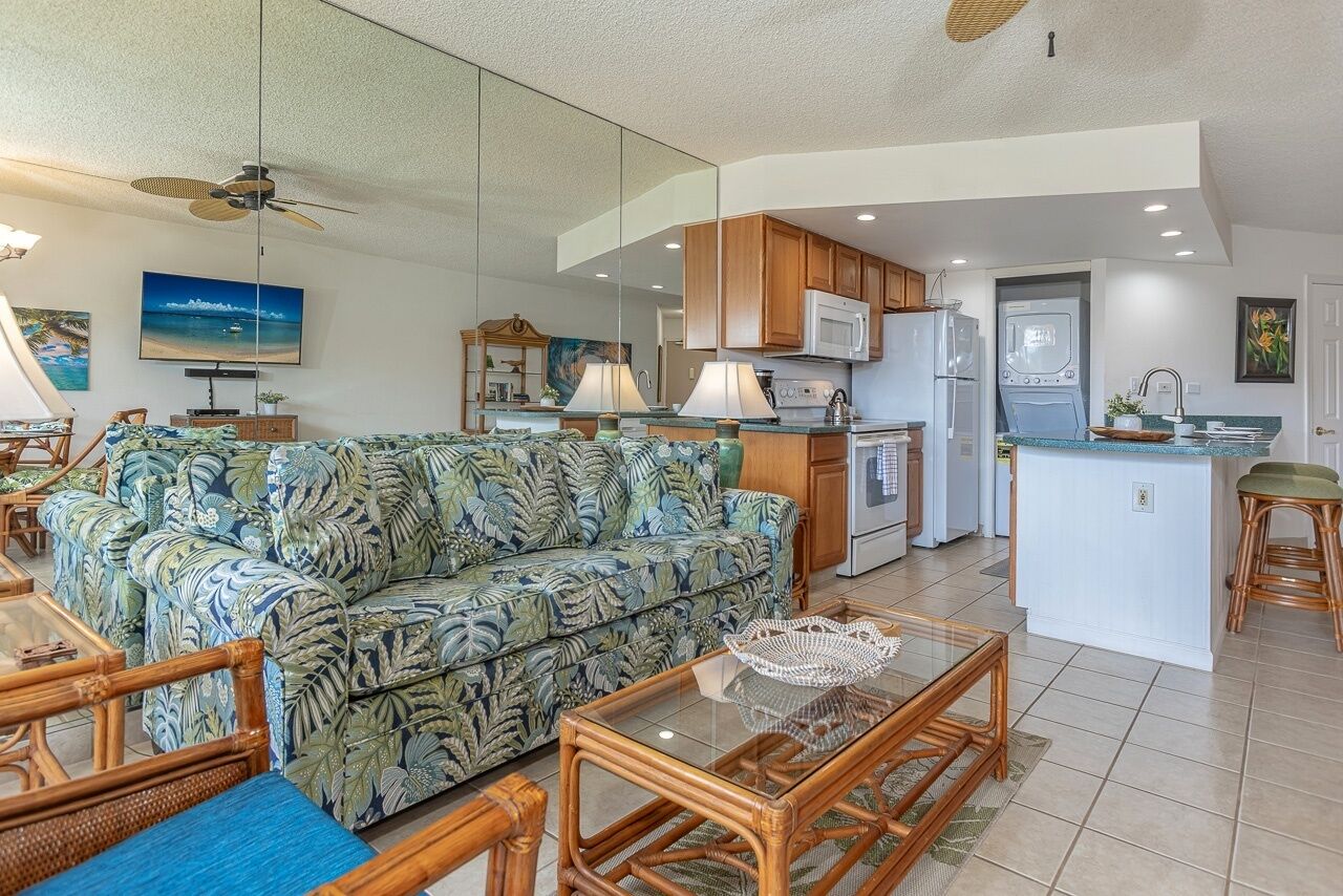 A modern living room with a sofa, coffee table, and dining area. It includes a kitchen with appliances and a large window.