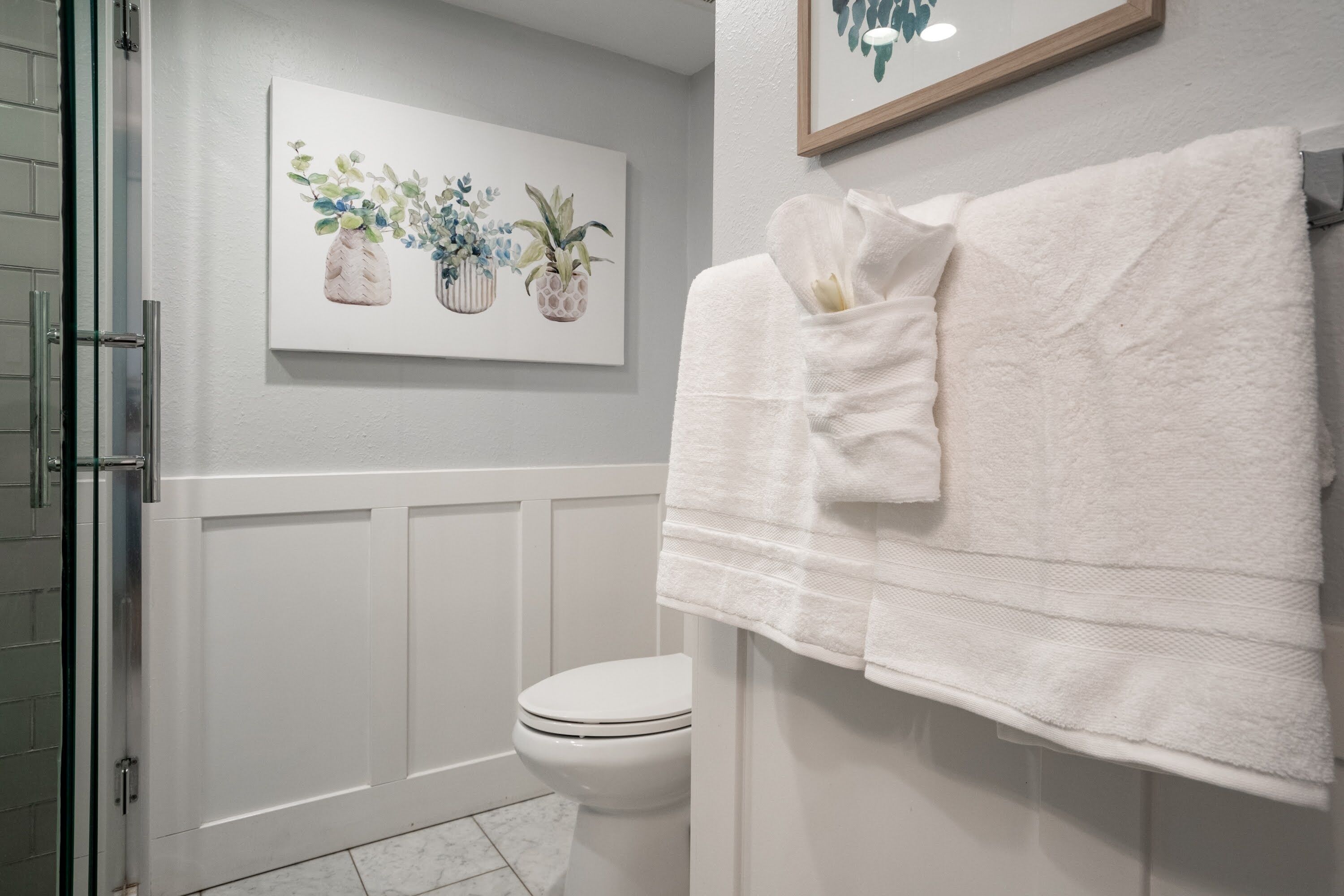 Une salle de bain avec des toilettes, un porte-serviettes blanc avec des serviettes, et une œuvre d’art encadrée représentant des plantes accrochée au mur.