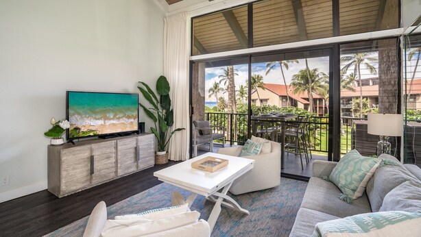 A spacious living area with a large TV, comfortable seating, and a balcony with a view of palm trees and buildings.
