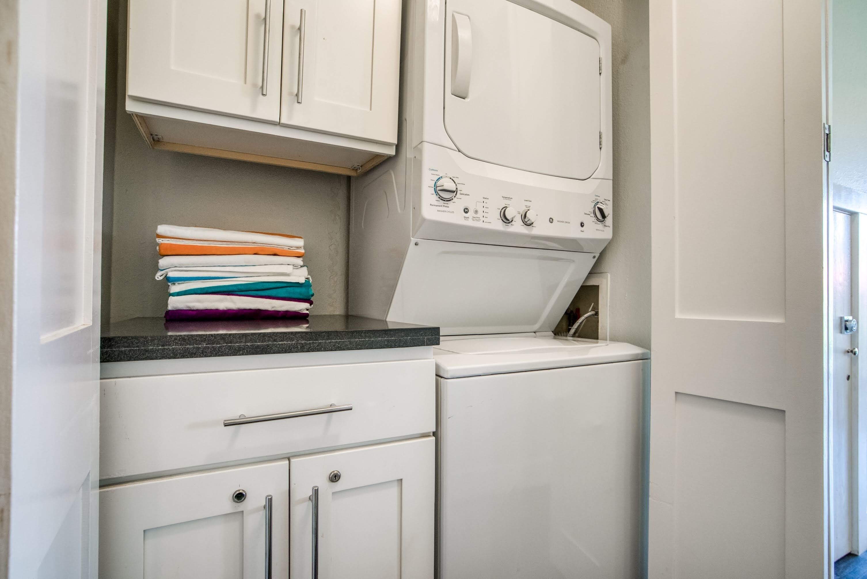 Un espace buanderie comprenant une machine à laver et un sèche-linge superposés, un plan de travail et un meuble contenant des vêtements pliés.