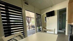 A room with a white table and chairs, a TV mounted on the wall, and a sliding door leading to an outdoor area with a patio and a view of a building. - D&I Mobile Home (Privlaka)