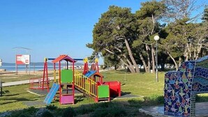 A playground with colorful equipment and a mosaic bench, set against a backdrop of trees and a clear sky. - D&I Mobile Home (Privlaka)