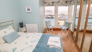 A bedroom with a bed, a radiator, a desk with a lamp, a chair, a window with a view of buildings, and a mirror. - Gower View (Tenby)