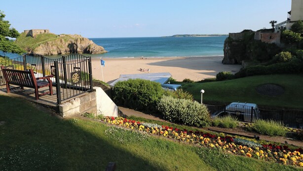 A coastal view with a beach, a garden with colorful flowers, and a historic building in the background. - Gower View (Tenby)