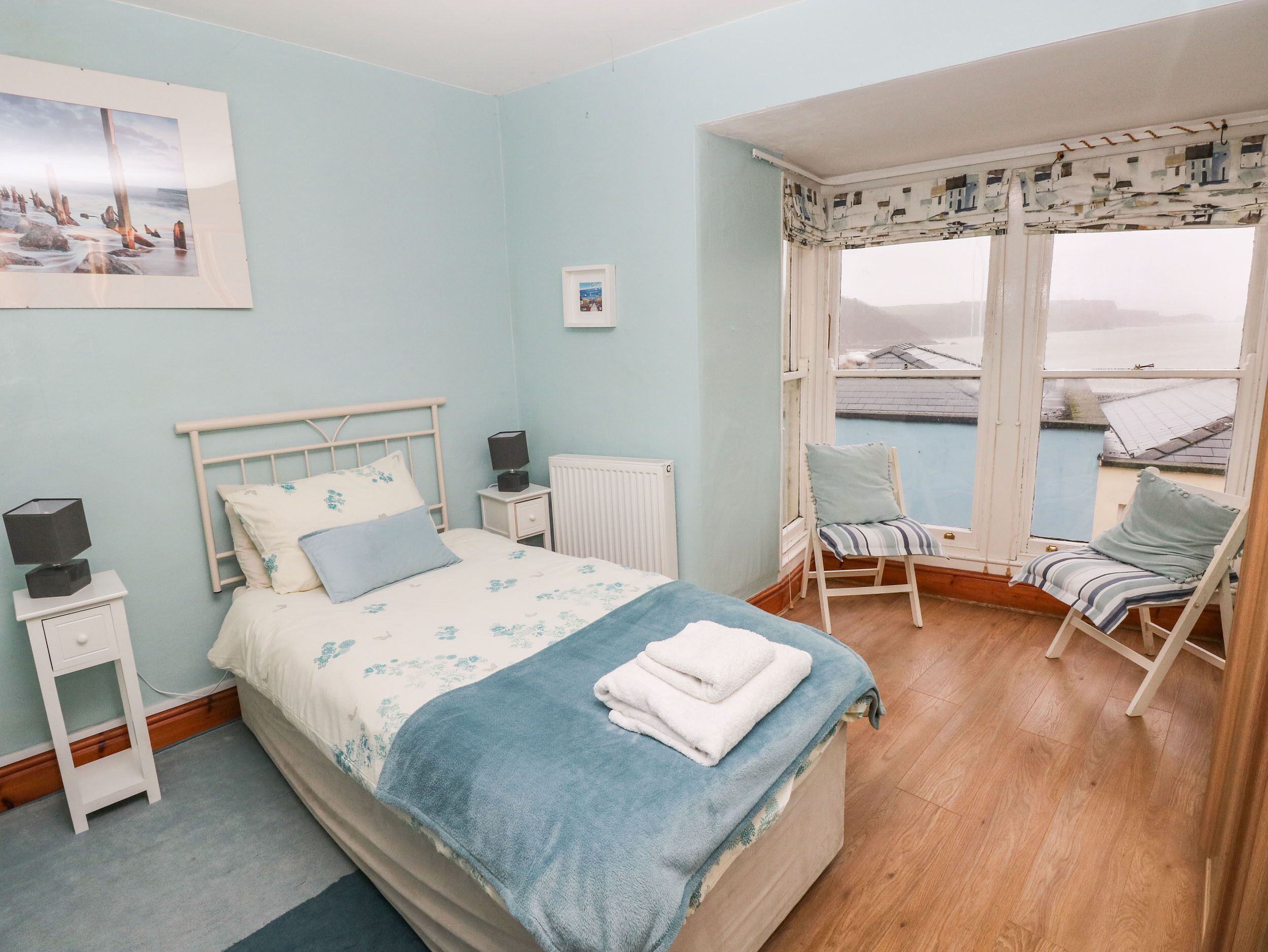 A bedroom with a bed, a nightstand, a chair by the window, and a view of the sea.