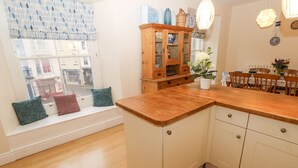 A kitchen with a wooden countertop, white cabinets, a window with a view of buildings, and a dining area with chairs and a table. - Gower View (Tenby)