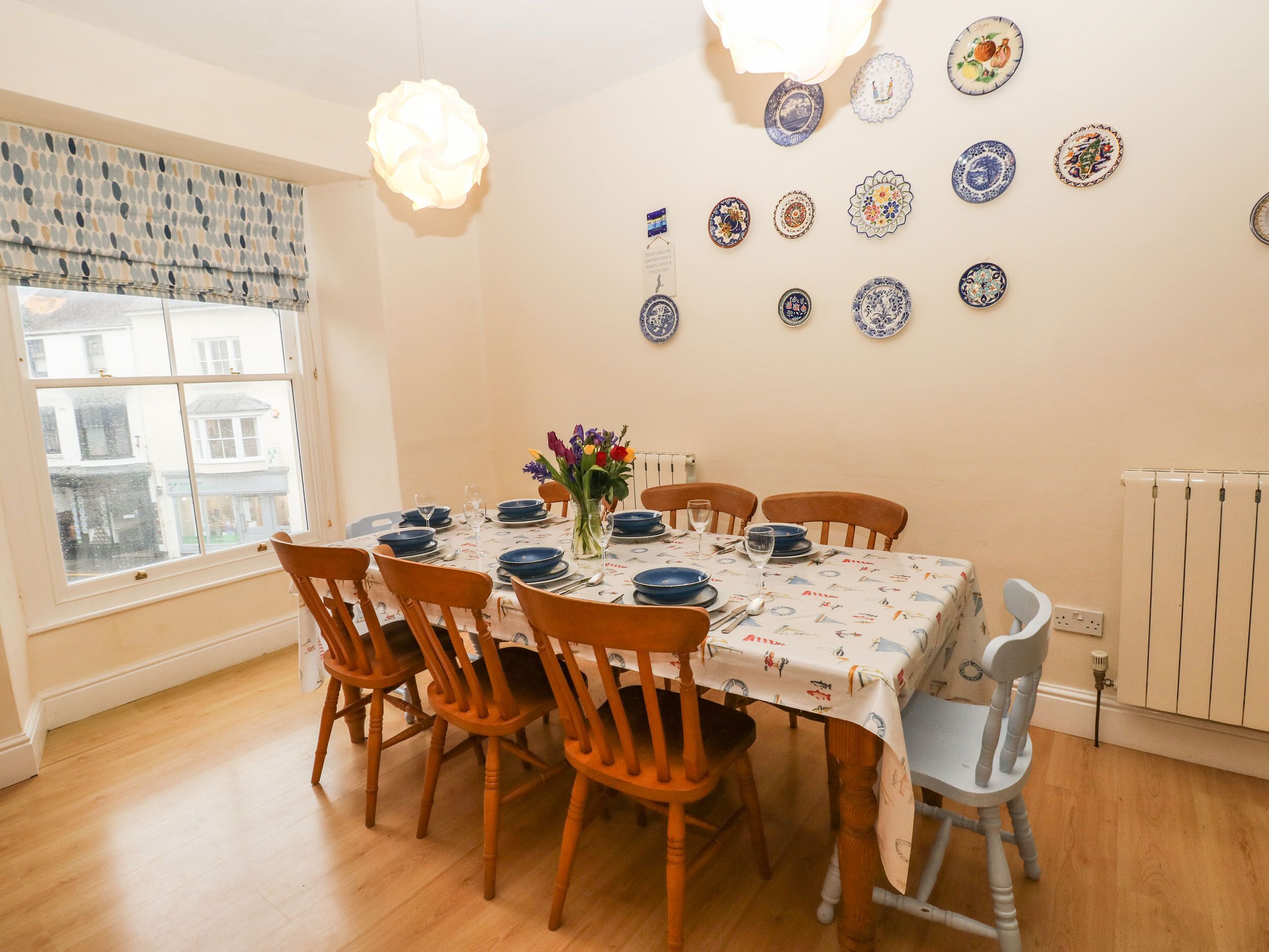 A dining room with a table set for six, surrounded by wooden chairs. The table is covered with a tablecloth featuring a nautical theme.