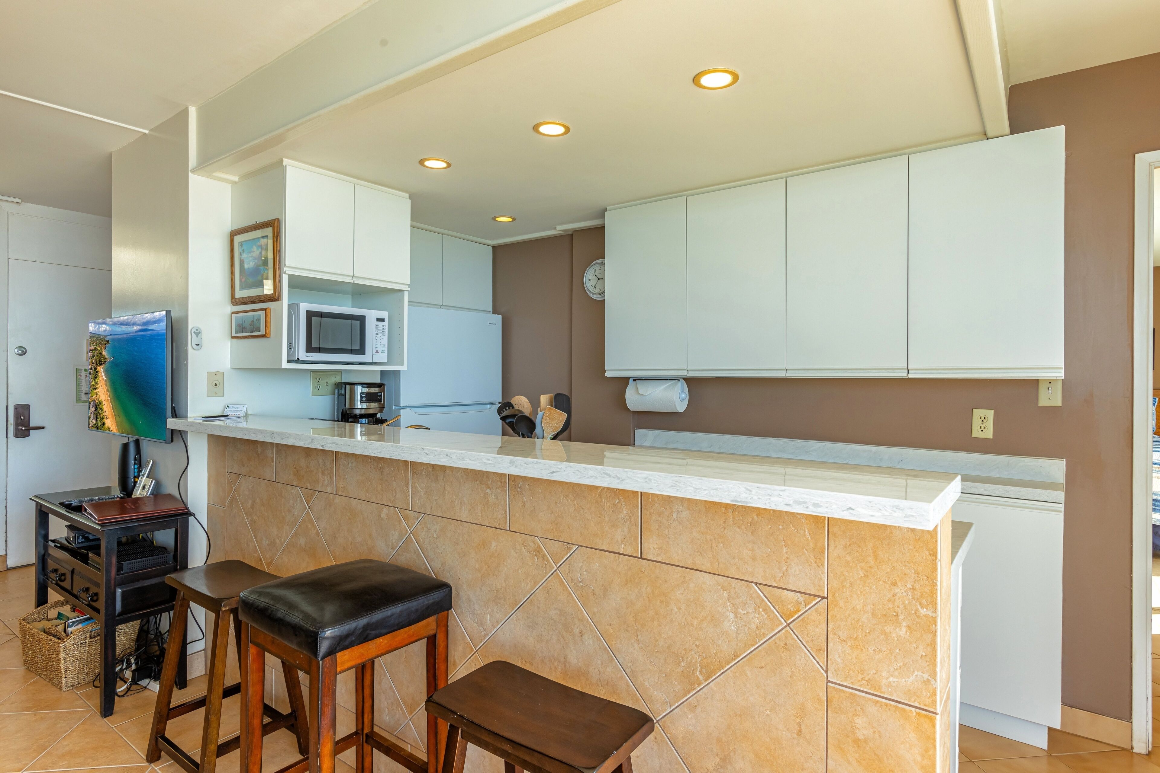 Cocina con gabinetes blancos, microondas, cafetera y un reloj en la pared. Hay dos taburetes y una mesa pequeña con una computadora portátil y una cesta.