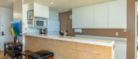 Cocina con gabinetes blancos, microondas, cafetera y un reloj en la pared. Hay dos taburetes y una mesa pequeña con una computadora portátil y una cesta.