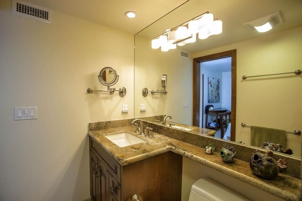 A bathroom with a double sink, a mirror with lights, a towel rack, and a toilet.