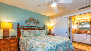 A bedroom with a bed, two bedside tables, a ceiling fan, and a bathroom with a vanity and mirror. - Sugar Beach Resort, #121 1 Bedroom Condo by RedAwning (Kihei)