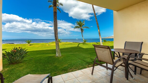 A patio with a table and chairs, overlooking a grassy area with palm trees and a view of the ocean. - Sugar Beach Resort, #121 1 Bedroom Condo by RedAwning (Kihei)