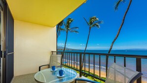 Un balcon avec une table et des chaises en verre, donnant sur une plage bordée de palmiers et sur l’océan.