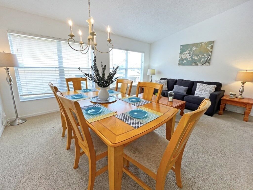 A dining area with a wooden table set for six, surrounded by chairs, a chandelier, and a vase with greenery. A sofa and a lamp are visible in the background.