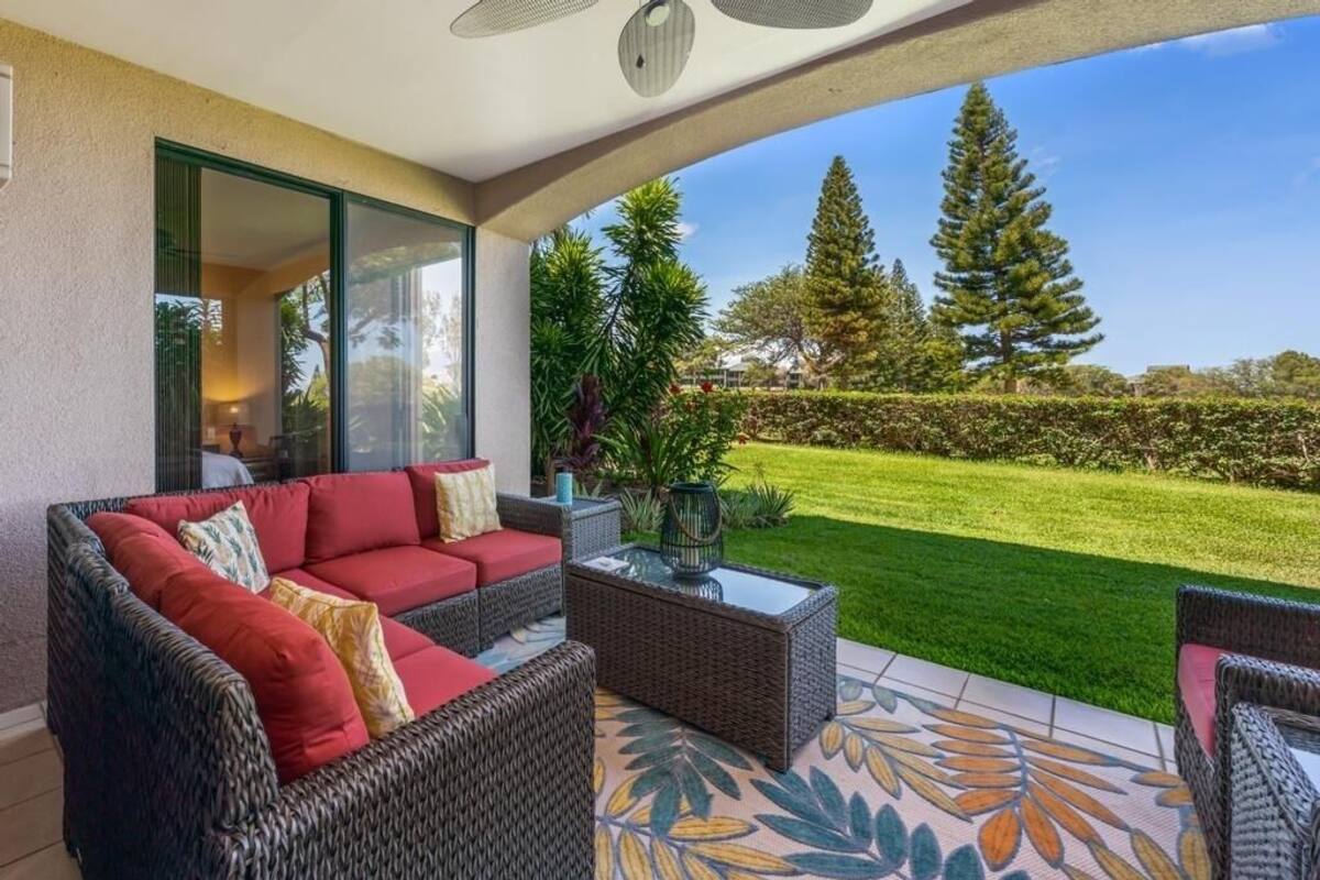 A covered patio area with wicker furniture, a glass table, and a view of a well-maintained lawn and trees.