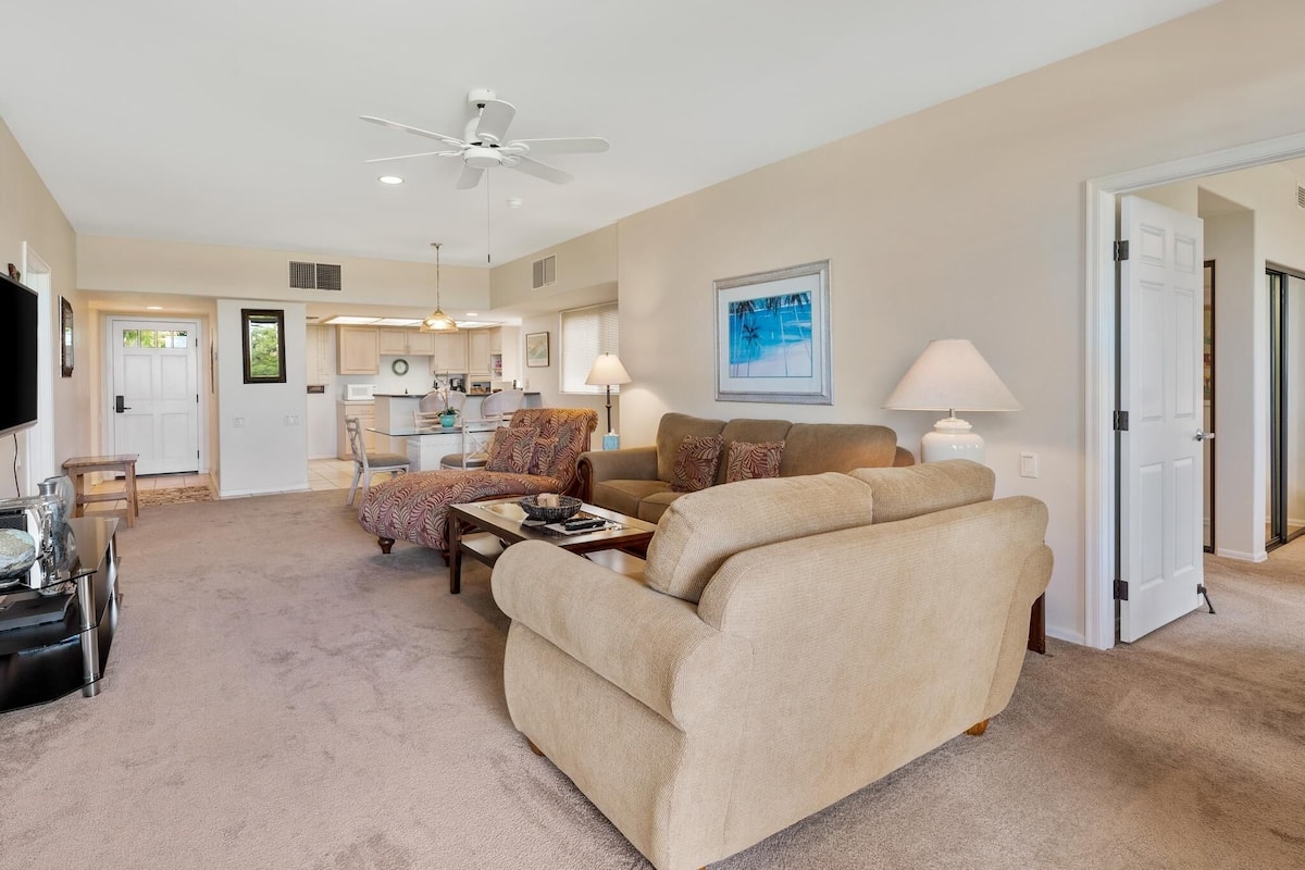 A spacious living room with a beige sofa, a coffee table, and a television. There is a kitchen area in the background with a dining table and chairs.