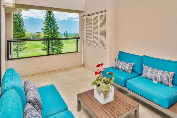 A balcony with a view of a golf course, a blue sofa set, and a wooden table with a plant. - Big Island Waikoloa Fairways by Coldwell Banker Island Vacations (Waikoloa)