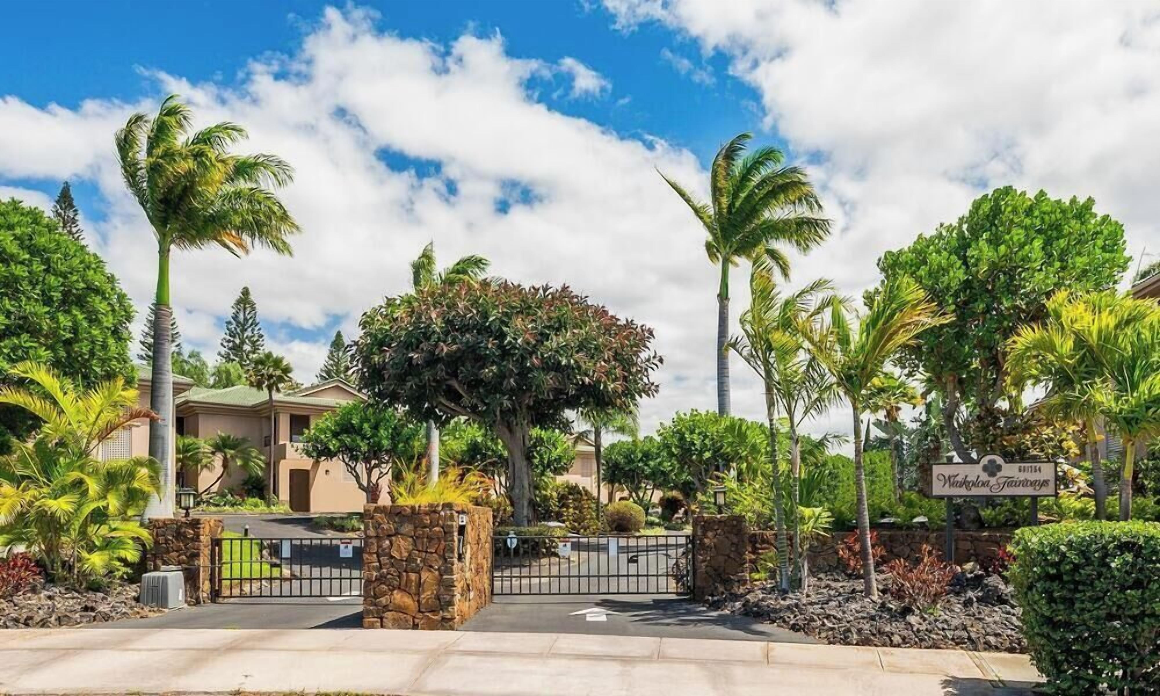 A gated entrance with a stone wall, palm trees, and a sign indicating a location.