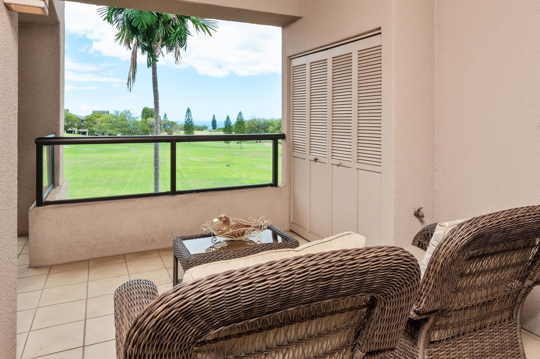 A balcony with wicker furniture, a glass table with a decorative item, and a view of a green field and palm tree.