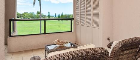 A balcony with wicker furniture, a glass table with a decorative item, and a view of a green field and palm tree.