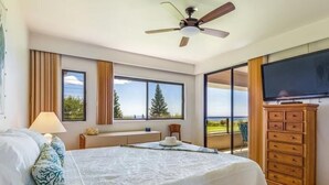 A bedroom with a bed, a ceiling fan, a TV, a dresser, and large windows offering a view of trees and a building. - Big Island Waikoloa Fairways by Coldwell Banker Island Vacations (Waikoloa)