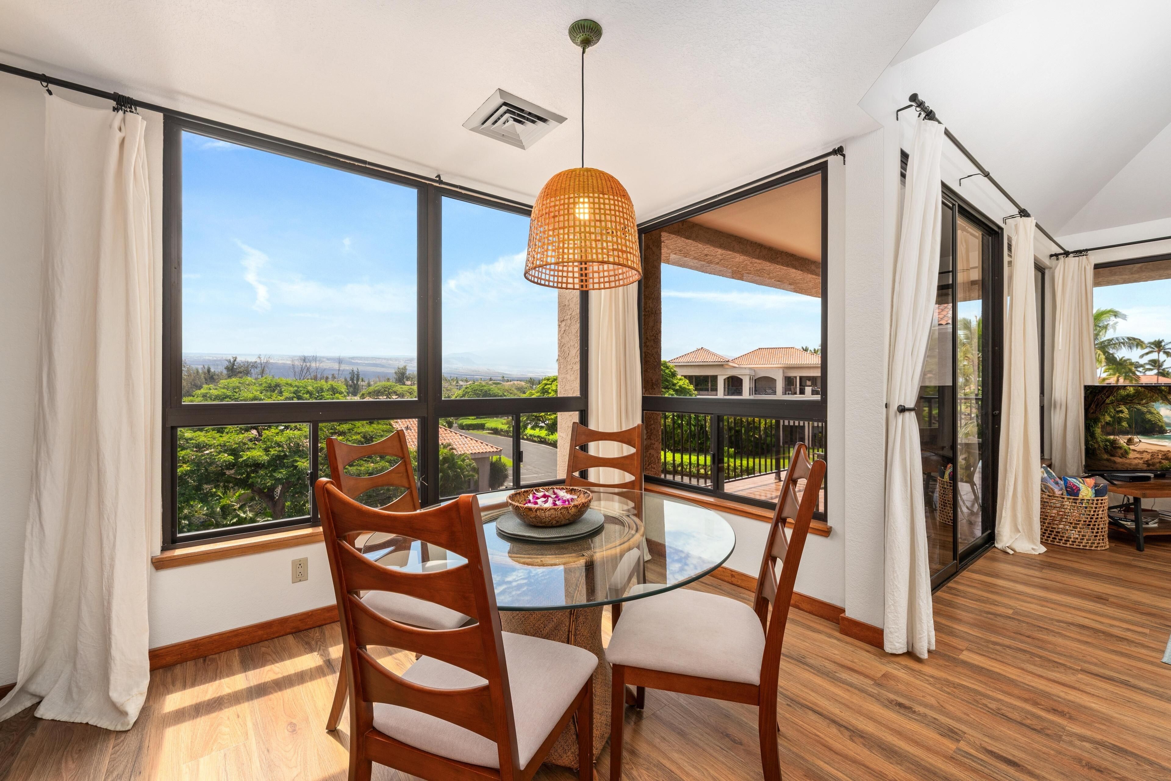 A dining area with a glass table and chairs, a woven pendant light, and a view of greenery and buildings through large windows.