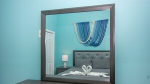 Ein Schlafzimmer mit einem großen Spiegel, einem Bett mit grauer Bettwäsche, einem Nachttisch mit Lampe und einem blauen Vorhang.