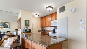 A kitchen with wooden cabinets, a granite countertop, a microwave, a stove, and a refrigerator. There is a dining area with wicker furniture and a table. - Sugar Beach Resort, #130 1 Bedroom Condo by RedAwning (Kihei)