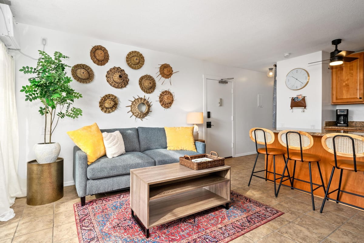 Ein Wohnzimmer mit grauem Sofa, gelben Kissen, einem Holztisch und einem gemusterten Teppich.