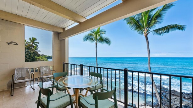 Varanda com mesa e cadeiras redondas, vista para uma praia com palmeiras e o oceano.
