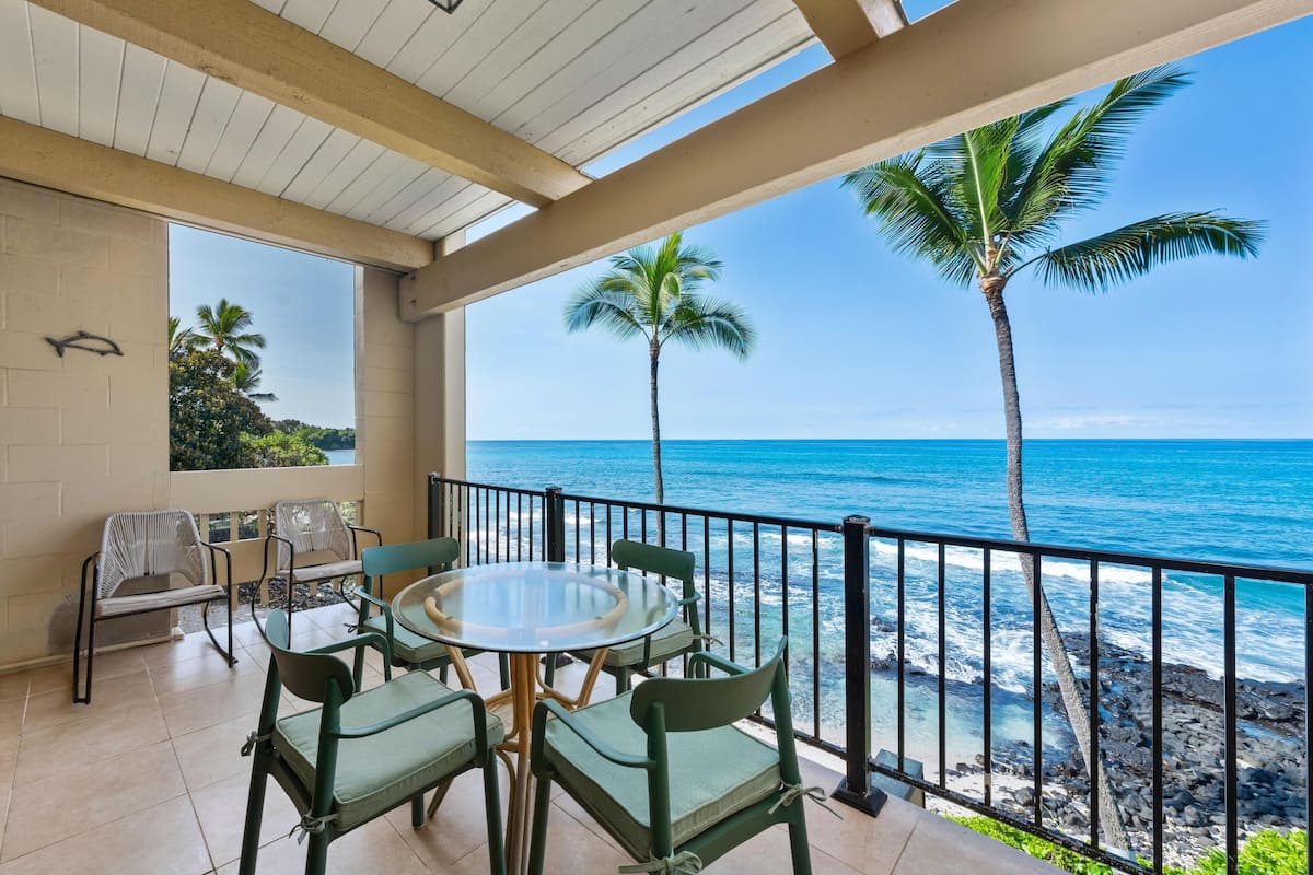 Ein Balkon mit rundem Tisch und Stühlen, der Blick auf einen Strand mit Palmen und den Ozean gerichtet ist.