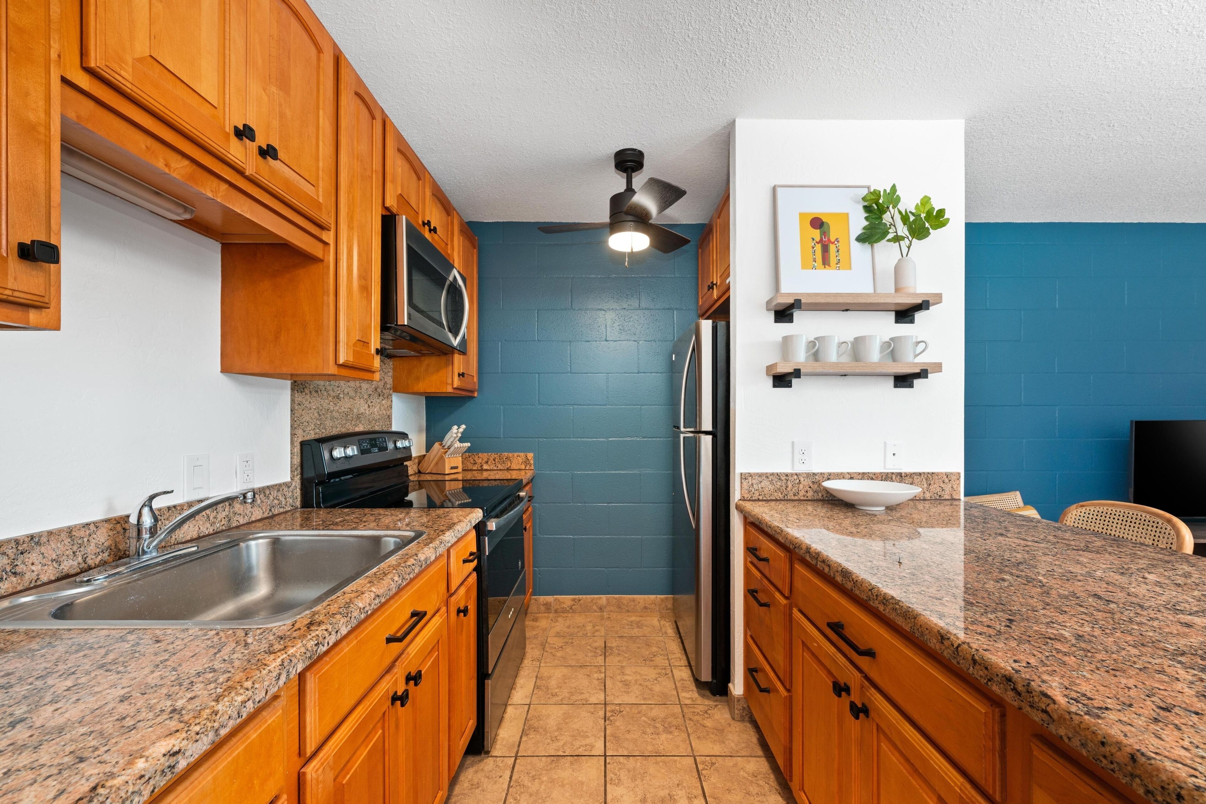 A kitchen with wooden cabinets, granite countertops, a stainless steel sink, a microwave, a refrigerator, and a ceiling fan.