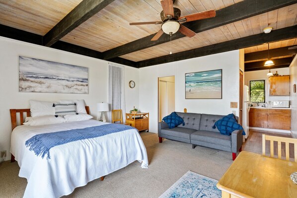 A bedroom with a bed, a gray sofa, a wooden ceiling fan, and a view of the beach through a window.