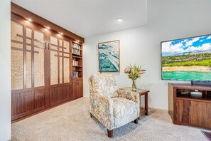 A living room with a large bookshelf, a comfortable armchair, a flat-screen TV, and a painting on the wall. - Wailea Elua, #0302 1 Bedroom Condo by RedAwning (Kihei)
