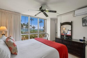 A hotel room with a bed, a ceiling fan, a large window with a view of palm trees and the ocean, a dresser, and a mirror. - Kamaole Nalu, #303 2 Bedroom Condo by RedAwning (Kihei)
