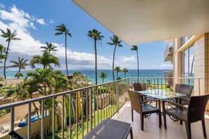 A balcony with a glass table and chairs, overlooking a pool and palm trees, with a view of the ocean and a building in the background. - Kamaole Nalu, #303 2 Bedroom Condo by RedAwning (Kihei)