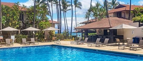 Ein Swimmingpool mit Liegestühlen und Sonnenschirmen, umgeben von Palmen und Gebäuden mit roten Ziegeldächern.