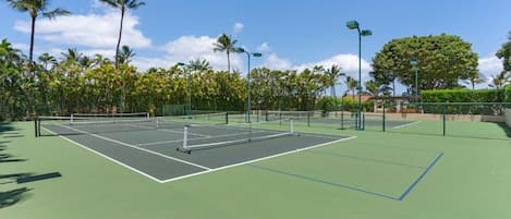 Ein Tennisplatz im Freien mit Netz, umgeben von Palmen und blauem Himmel.