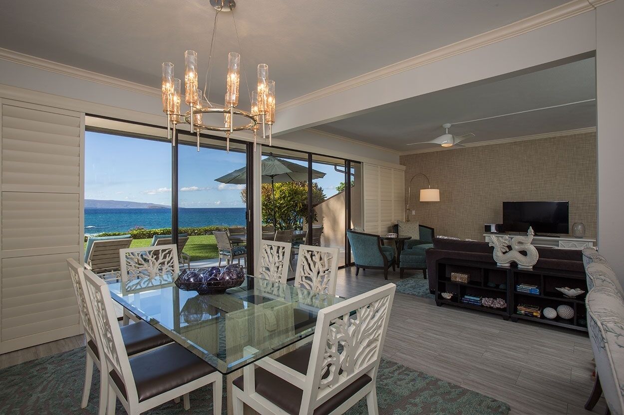 Uma sala de jantar com mesa e cadeiras de vidro, um candelabro e vista para o oceano.