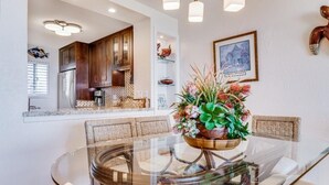 A modern kitchen with wooden cabinets, a built-in oven, and a dining area with a glass table and chairs. - Menehune Shores, #115^ 3 Bedroom Condo by RedAwning (Kihei)