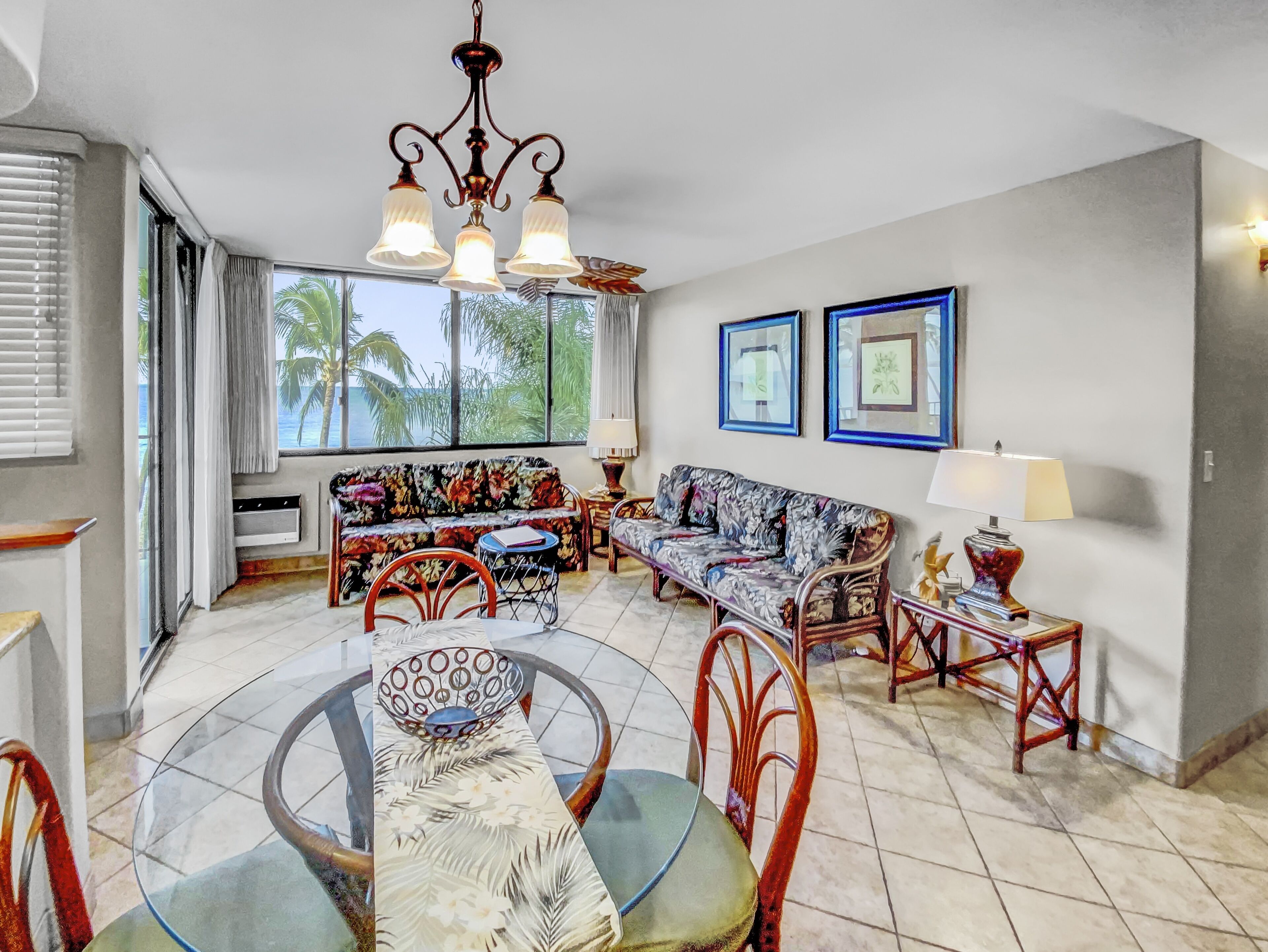 A spacious living room with a glass dining table, two sofas, a chandelier, and a view of palm trees through large windows.