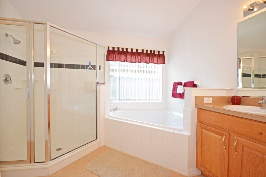 A bathroom with a glass shower enclosure, a bathtub, a vanity with a mirror, and a window with a red valance.