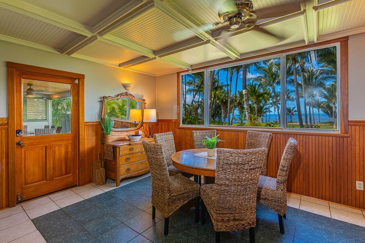 A dining area with a round table and wicker chairs, a wooden cabinet with drawers, a mirror, and a view of palm trees through large windows.