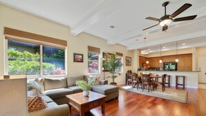A spacious living room with a wooden floor, a ceiling fan, and large windows offering a view of greenery outside. - Big Island Fairways At Mauna Lani 1705 3 Bedroom Townhouse (Kamuela)