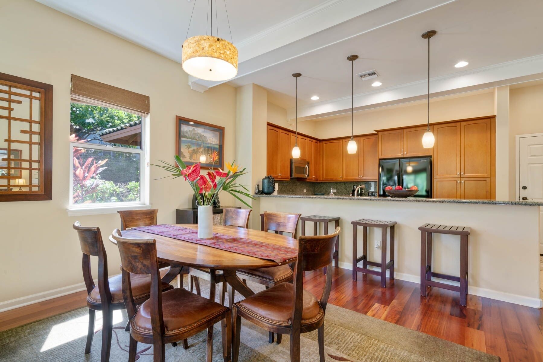 A dining area with a wooden table and chairs, a vase with flowers, and a kitchen with wooden cabinets and a refrigerator.