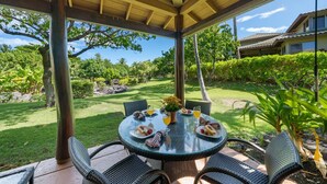 A covered outdoor dining area with a round table and wicker chairs, surrounded by lush greenery and a view of a house in the background. - Big Island Fairways At Mauna Lani 1705 3 Bedroom Townhouse (Kamuela)