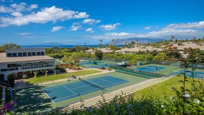 A tennis court complex with multiple courts, surrounded by lush greenery and palm trees, overlooking a scenic ocean view.