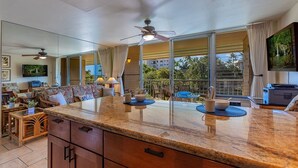 A modern kitchen with a granite countertop, wooden cabinets, and a dining area with wicker furniture. - Kihei Kai Nani by Coldwell Banker Island Vacations (Kihei)