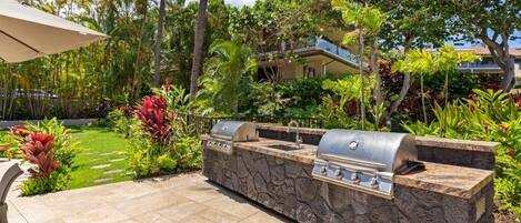 An outdoor barbecue area with stone countertops and two grills, surrounded by tropical plants and a clear sky.